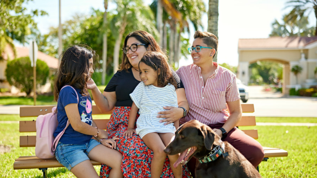 LGBTQ couple and children sitting on a bench in a park smiling with pet dog.