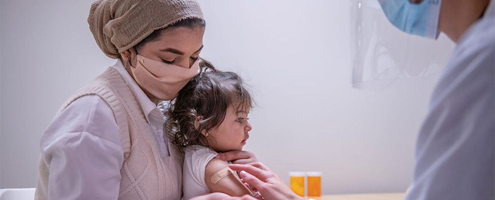 Photo of masked, young woman in doctor's office setting wearing headscarf, blouse and sweater vest holding young female child on lap. A female doctor with mask and face shield is applying band aid to child's upper arm.