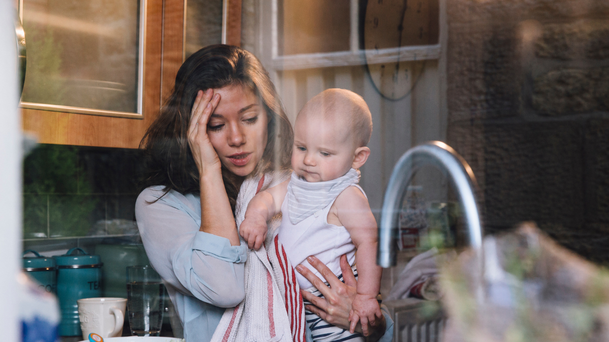 Young mother holding baby and looking overwhelmed in the kitchen
