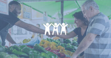 Vendor and woman shaking hands over bell pepper stand at outdoor market.