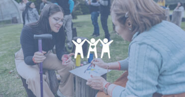 Two women painting on paper on top of stump at outdoor outreach event.